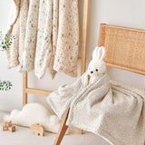 Baby floral gauze blanket with a bunny design draped over a chair in a room with hanging baby clothes and toys.
